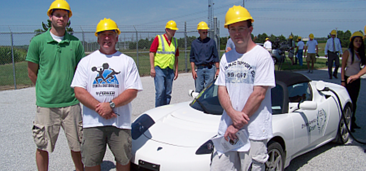 students and teachers standing in front of a car with hardhats on 